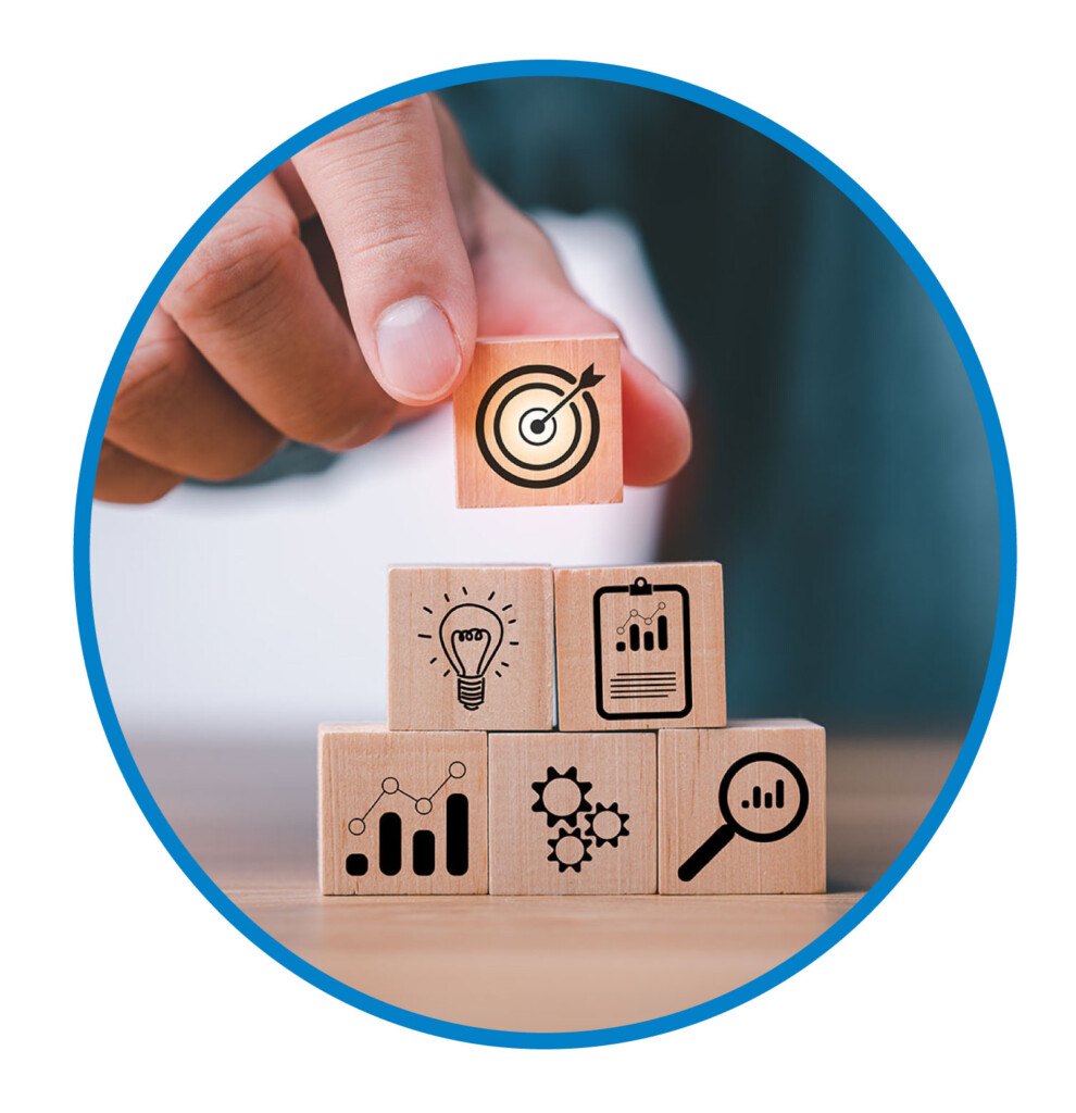 A hand stacks wooden blocks with icons of a target, chart, light bulb, clipboard, gears, and magnifying glass, symbolizing goals, strategy, innovation, research, and analysis.