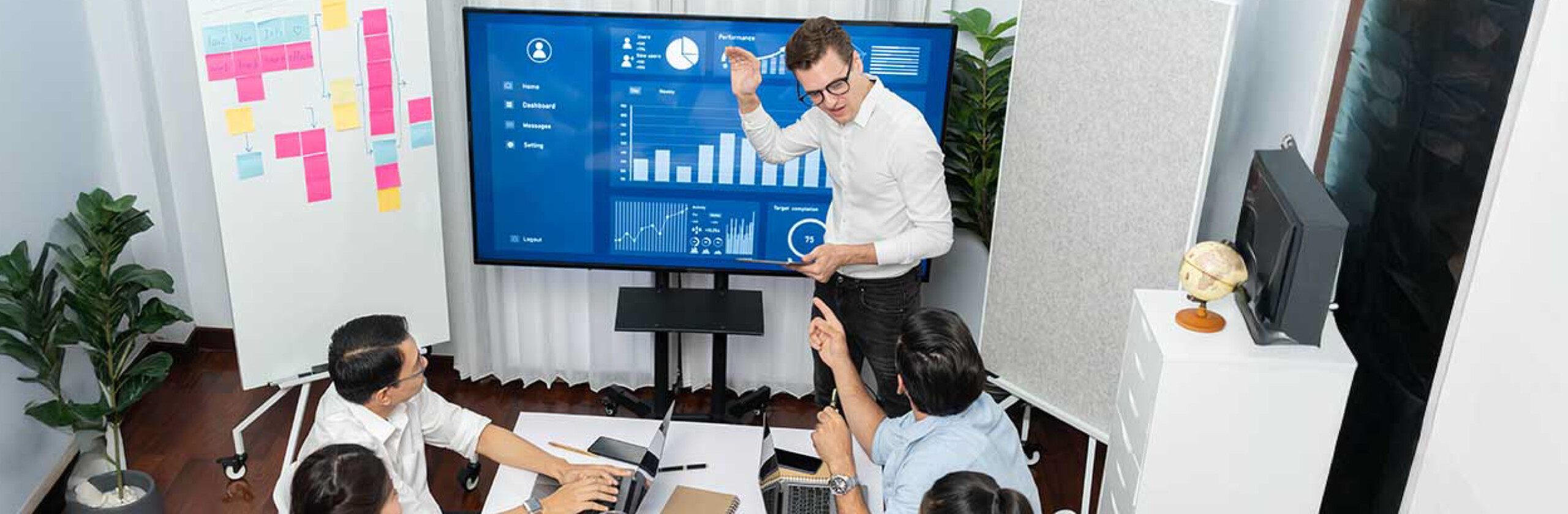 Image- What are the 5 Stages of Project Management? Team gathered in a conference room while a presenter explains project data on a screen with charts and graphs.